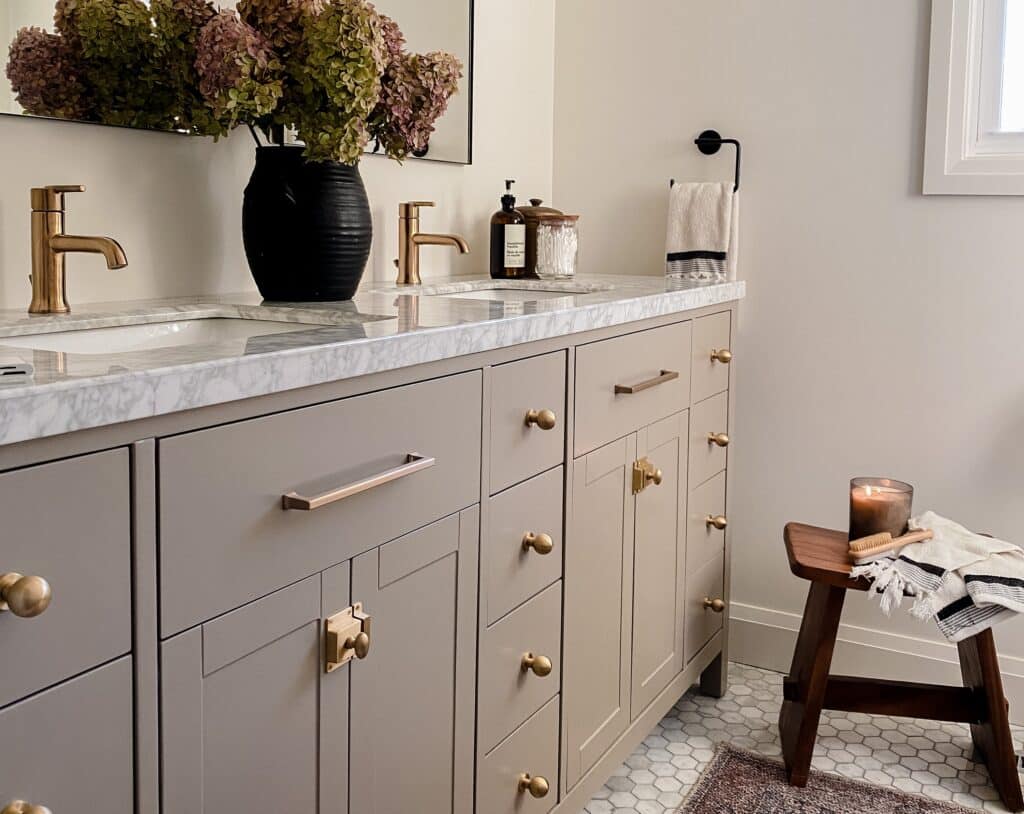 Modern bathroom with dual sinks, brass fixtures, marble countertop, textured cabinets, vase with flowers, and lit candle on wooden stool.