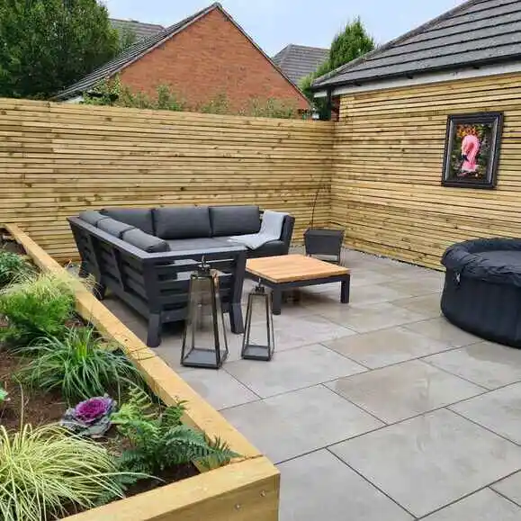 low-maintenance-porcelain-patio-paving-slabs-uk Modern backyard patio with gray sectional sofa, wooden table, lanterns, and Jacuzzi, surrounded by a wooden fence and greenery. | Sky Rye Design Modern backyard patio with gray sectional sofa, wooden table, lanterns, and Jacuzzi, surrounded by a wooden fence and greenery.
