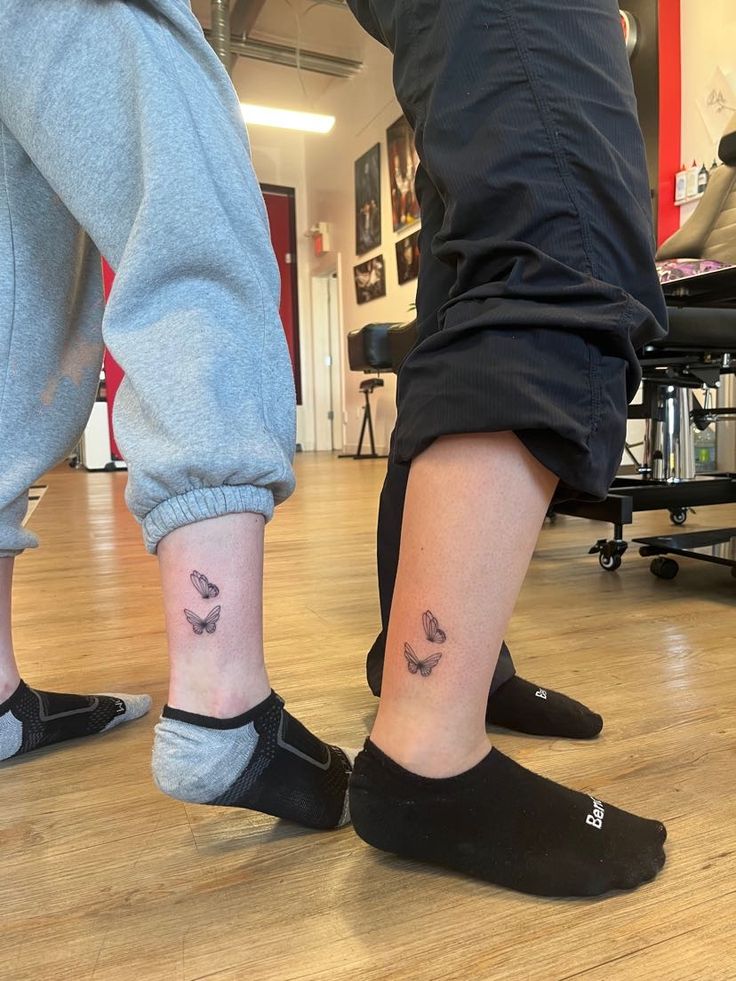 matching butterflies🤍 Two people in joggers showcase small butterfly tattoos on their ankles inside a tattoo studio, highlighting personal expression. | Sky Rye Design Two people in joggers showcase small butterfly tattoos on their ankles inside a tattoo studio, highlighting personal expression.