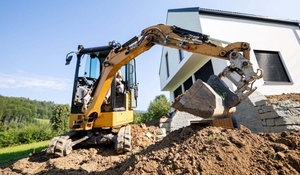 mini-excavator-min Yellow excavator digging soil next to a modern house on a sunny day, with a green landscape in the background. | Sky Rye Design Yellow excavator digging soil next to a modern house on a sunny day, with a green landscape in the background.