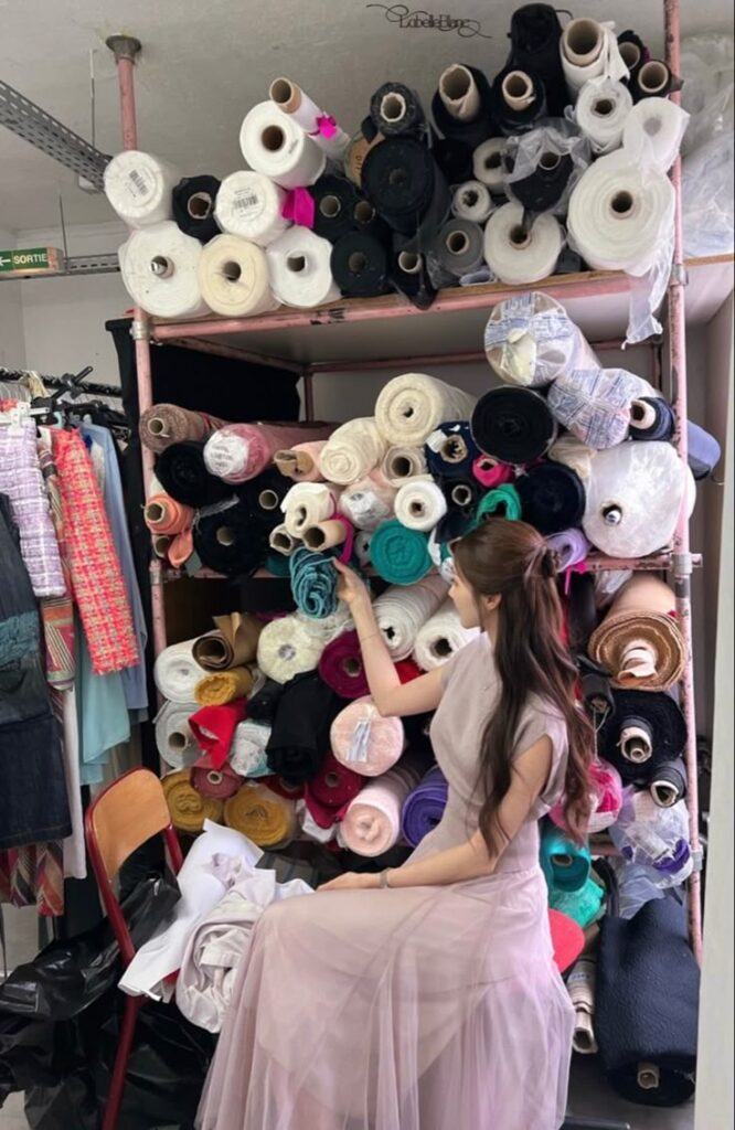 nehwa Woman in a pink dress selecting fabric rolls from a shelf in a fashion studio. Colorful textiles in the background. | Sky Rye Design Woman in a pink dress selecting fabric rolls from a shelf in a fashion studio. Colorful textiles in the background.