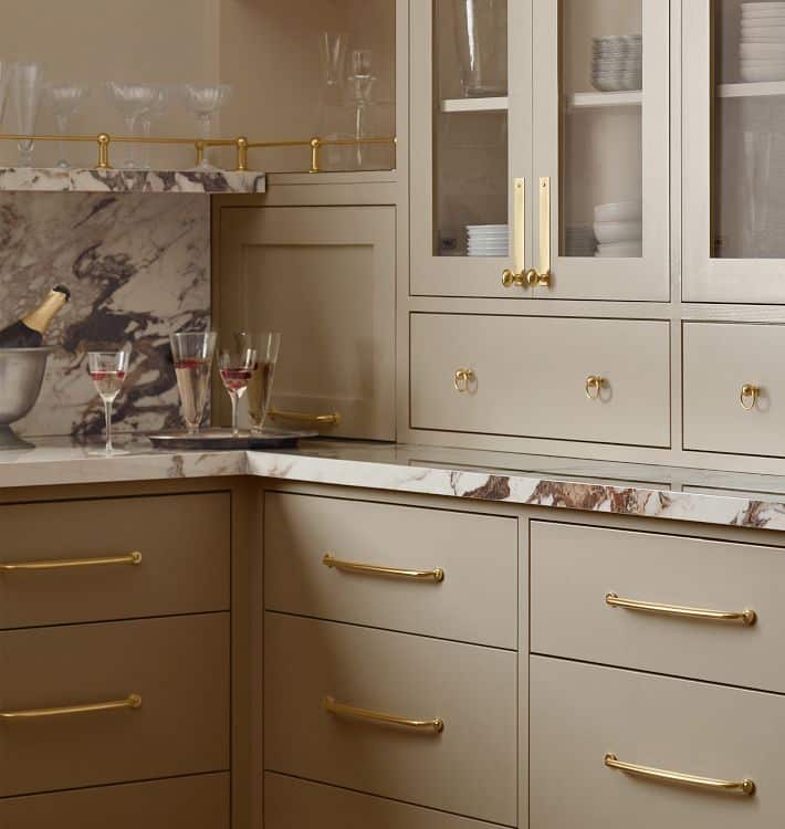 Elegant beige kitchen with marble countertops, gold handles, and glass cabinets, featuring wine glasses and a champagne bottle on display.