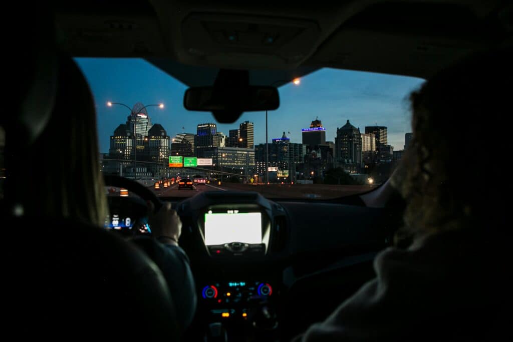 pexels-wendywei-1179579 City skyline visible from a car at dusk, with two people in the front seats and illuminated buildings ahead. Urban evening drive. | Sky Rye Design City skyline visible from a car at dusk, with two people in the front seats and illuminated buildings ahead. Urban evening drive.