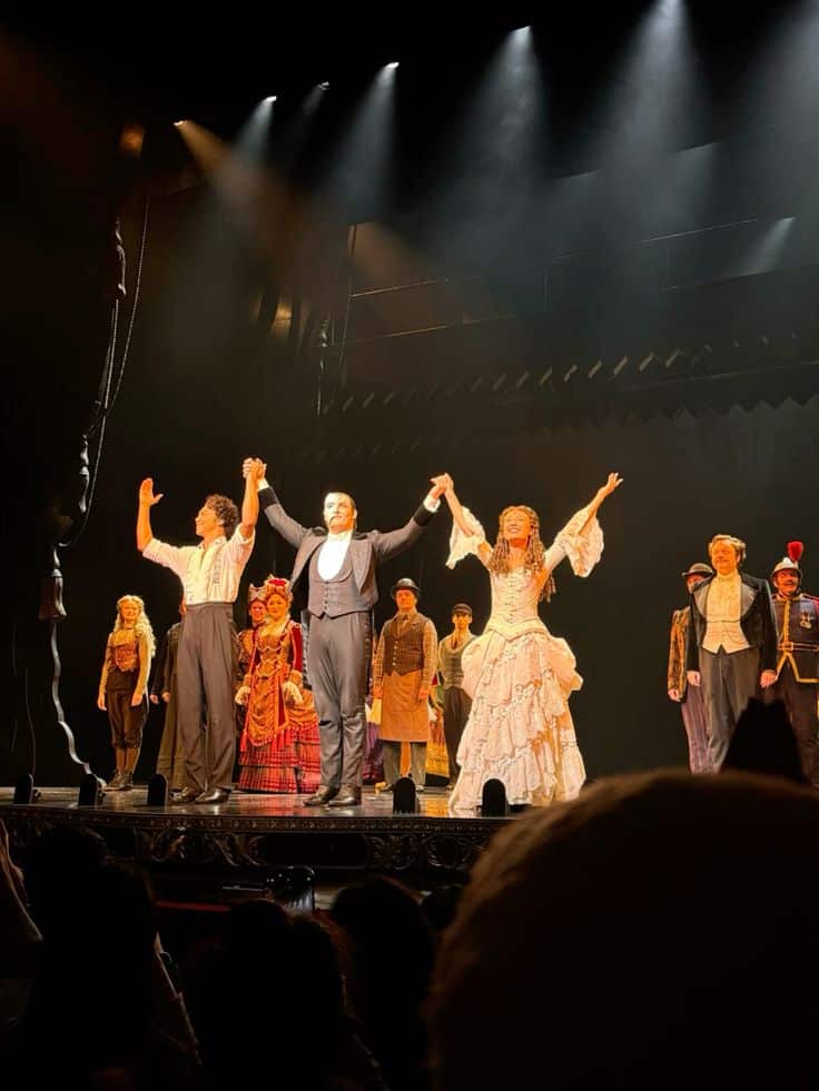 phantom of the opera london Theater cast taking a bow on stage in spotlight, wearing elaborate costumes, with audience visible in foreground. | Sky Rye Design Theater cast taking a bow on stage in spotlight, wearing elaborate costumes, with audience visible in foreground.