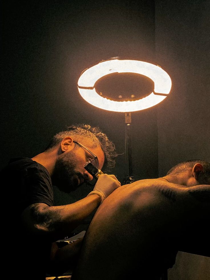 photography_ Tattoo artist focused on creating detailed ink design under circular light in a dimly lit studio. | Sky Rye Design Tattoo artist focused on creating detailed ink design under circular light in a dimly lit studio.