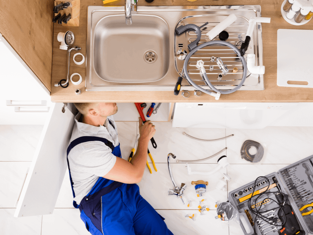 plumbing-services-types Plumber in blue overalls fixing a sink with tools spread around, showcasing a repair in progress under a kitchen counter. | Sky Rye Design Plumber in blue overalls fixing a sink with tools spread around, showcasing a repair in progress under a kitchen counter.