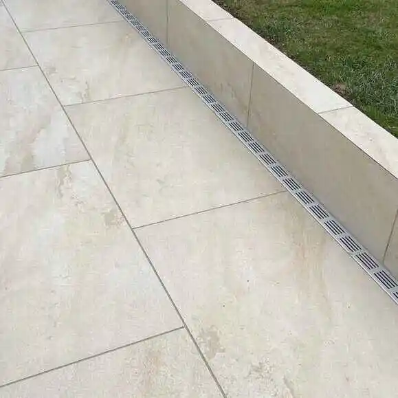 porcelain-patio-drainage-channel-installation-uk Cream stone patio with linear drainage alongside a small grass lawn, offering modern outdoor design and efficient water management. | Sky Rye Design Cream stone patio with linear drainage alongside a small grass lawn, offering modern outdoor design and efficient water management.