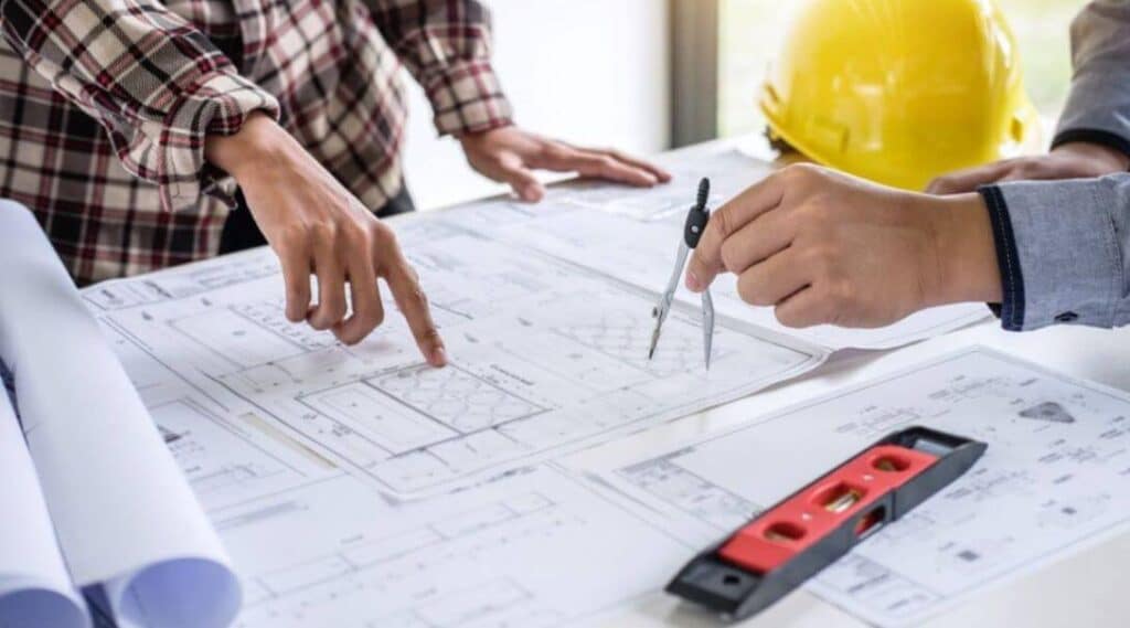 pre-construction Architects reviewing blueprints with compass and level on table, hard hat in background. | Sky Rye Design Architects reviewing blueprints with compass and level on table, hard hat in background.