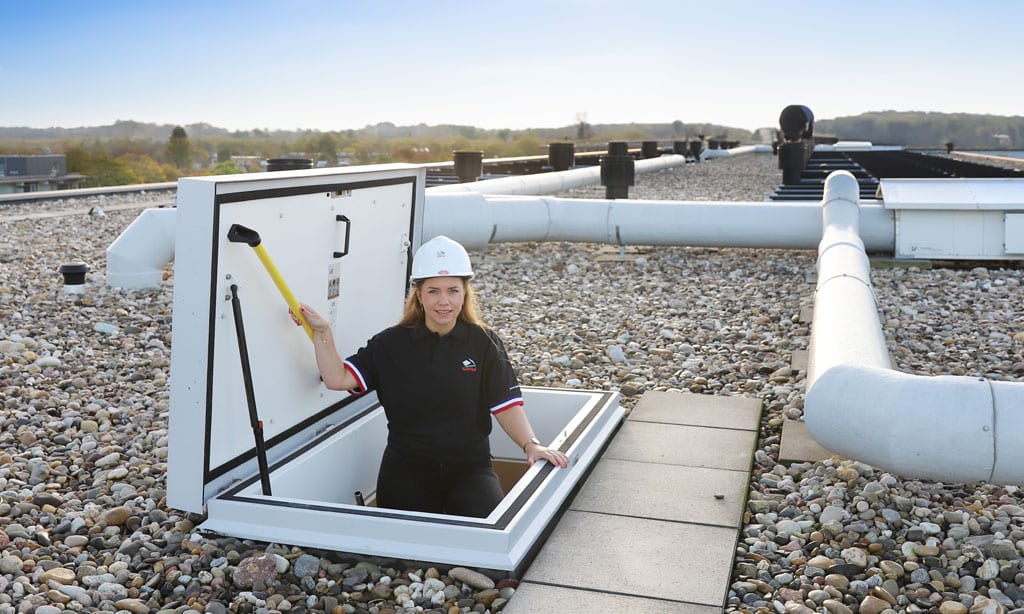 Person in a helmet emerging from a rooftop hatch on a gravel-covered roof, holding a tool. Pipes and landscape seen in the background.