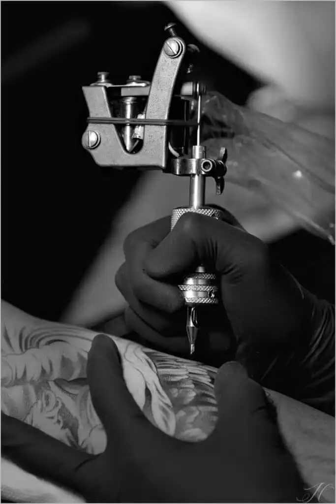 Tattoo artist in gloves using a tattoo machine for intricate black and white design on client's arm.