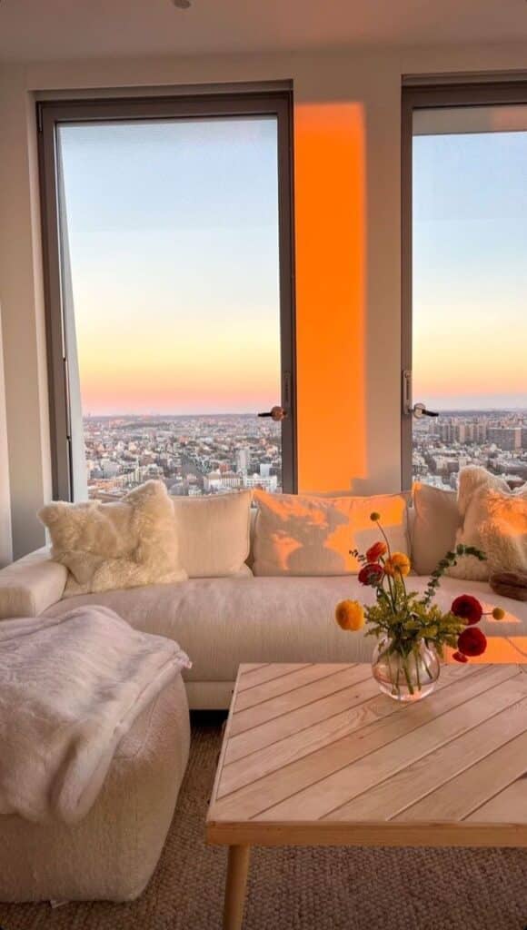 Cozy living room with a white sofa, colorful flowers on a wooden table, and a city view at sunset through large windows.