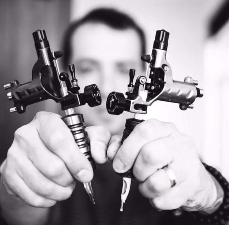 tattoo Tattoo artist holding two detailed tattoo machines, ready for work. Black and white image highlighting the equipment's precision. | Sky Rye Design Tattoo artist holding two detailed tattoo machines, ready for work. Black and white image highlighting the equipment's precision.