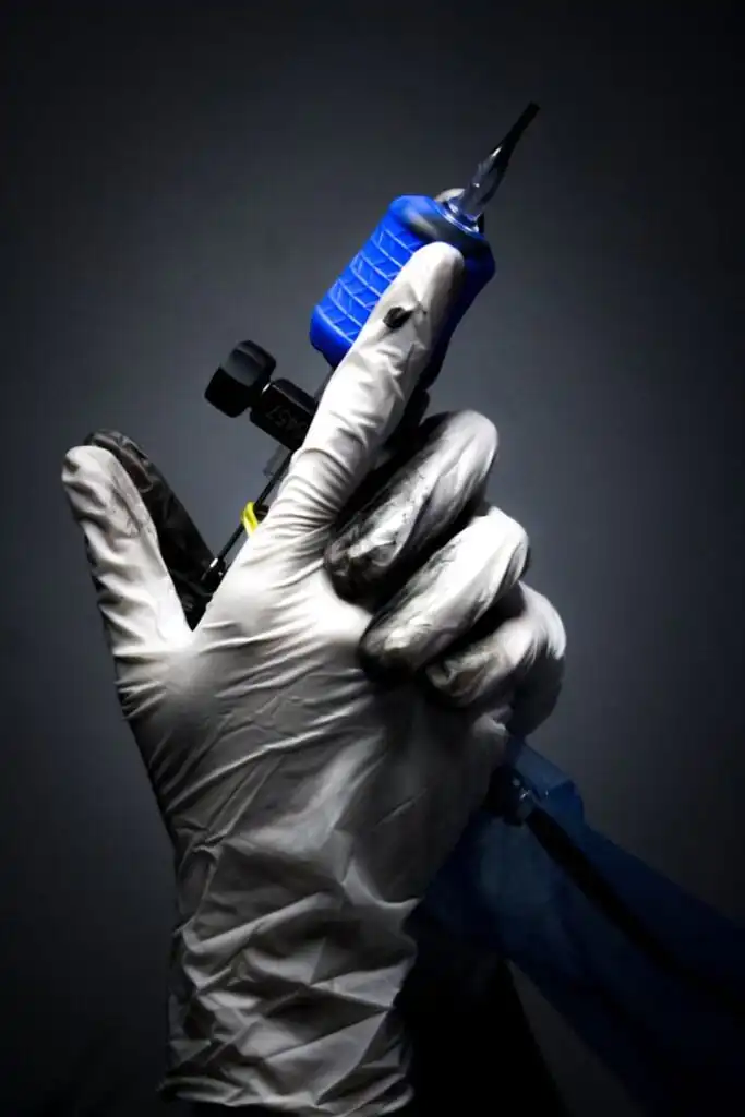 Gloved hand holding a tattoo machine with a blue grip, ready for artistic tattooing on a dark background.