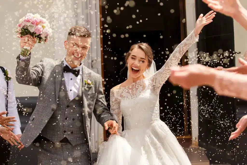 Joyful newlyweds celebrate wedding exit amid confetti shower, bride in elegant lace gown and groom in stylish grey suit, holding bouquet.