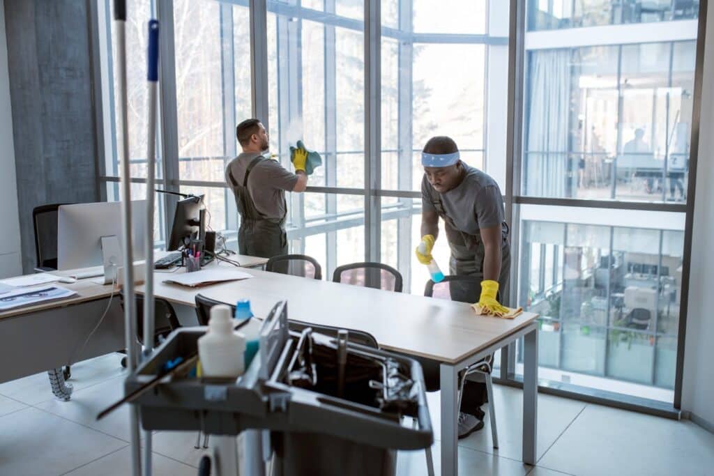 Office cleaning crew sanitizing desks and windows with supplies cart nearby, ensuring a spotless work environment in a modern building.