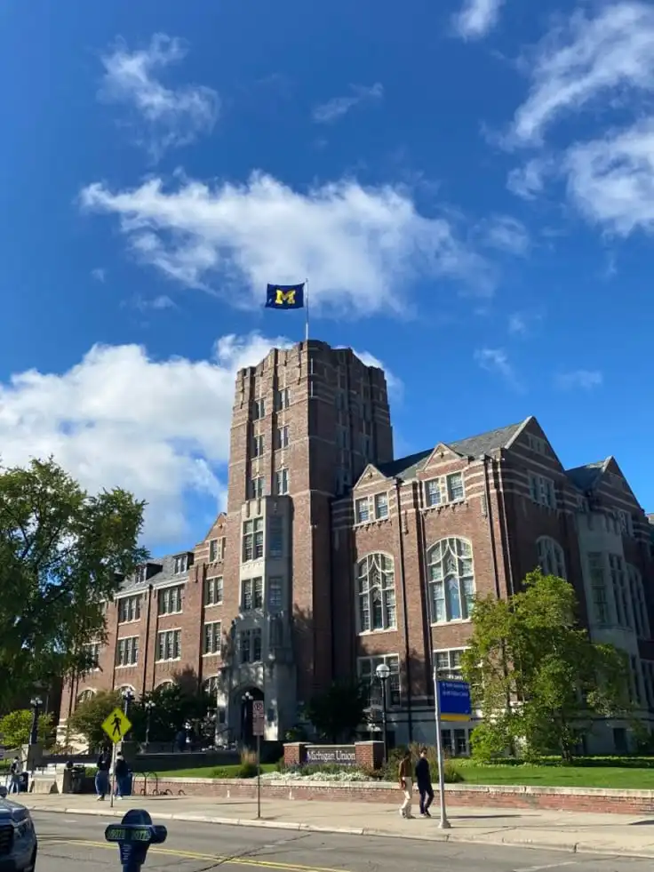 Red-brick building with a flag waving on top under a clear blue sky at Michigan Union, Ann Arbor, surrounded by trees and pedestrians.