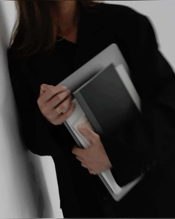 _ Person in black holding notebooks and a laptop, leaning against a wall, ring on finger. Stylish and professional, blurred motion effect. | Sky Rye Design Person in black holding notebooks and a laptop, leaning against a wall, ring on finger. Stylish and professional, blurred motion effect.