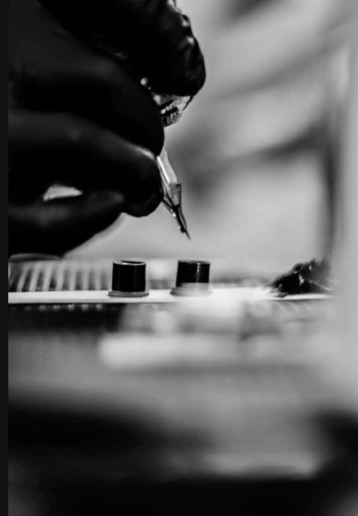 _ Black and white image of a tattoo artist's gloved hand holding a tattoo machine, with ink cups in the foreground. | Sky Rye Design Black and white image of a tattoo artist's gloved hand holding a tattoo machine, with ink cups in the foreground.