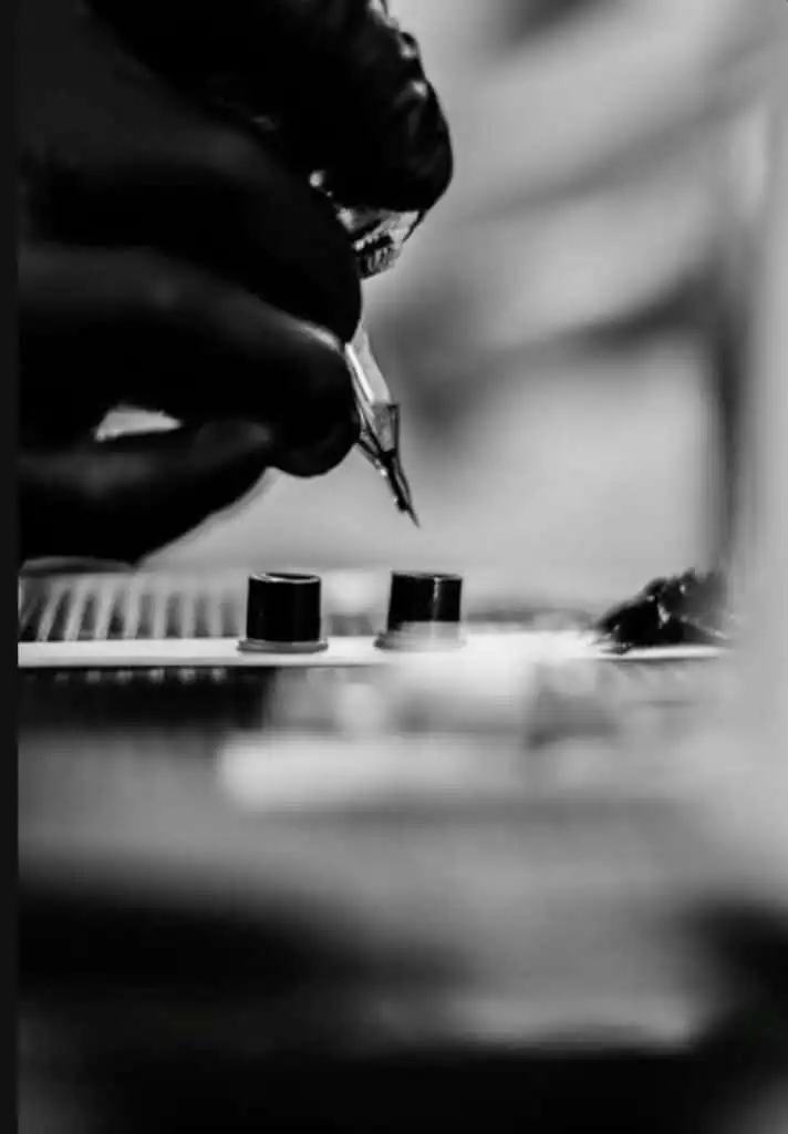 Black and white image of a tattoo artist's gloved hand holding a tattoo machine, with ink cups in the foreground.
