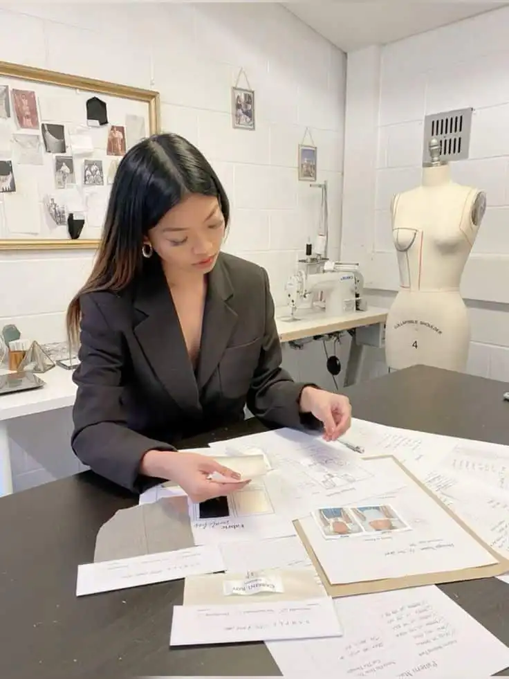 _ Fashion designer reviewing fabric samples and sketches in a creative studio with a dress form and sewing machine in the background. | Sky Rye Design Fashion designer reviewing fabric samples and sketches in a creative studio with a dress form and sewing machine in the background.
