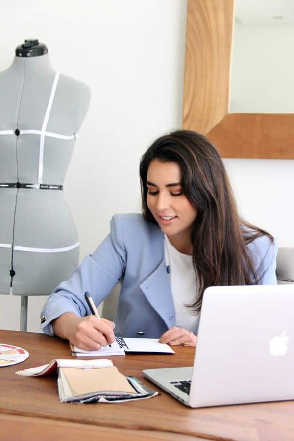 Fashion designer sketching ideas at desk with laptop, mannequin, and fabric swatches. Creativity and design process in a modern workspace.