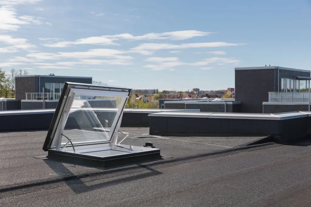 velux-glass-rooflight-electric-exit-roof-windows-archello17570614114412 Open rooftop hatch under clear blue sky, providing access and ventilation to modern building, surrounded by cityscape. | Sky Rye Design Open rooftop hatch under clear blue sky, providing access and ventilation to modern building, surrounded by cityscape.