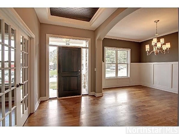 wainscotting Spacious foyer with wooden floors, elegant wainscoting, chandelier, and an open front door leading to a sunlit exterior view. | Sky Rye Design Spacious foyer with wooden floors, elegant wainscoting, chandelier, and an open front door leading to a sunlit exterior view.