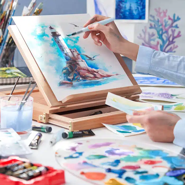 watercolour-paint-12-colours-12-x-12-ml-0-74-us-fl-oz_fqX3vCao_700x700 Artist painting a vibrant lighthouse scene with watercolors on an easel, surrounded by art supplies and colorful palettes. | Sky Rye Design Artist painting a vibrant lighthouse scene with watercolors on an easel, surrounded by art supplies and colorful palettes.