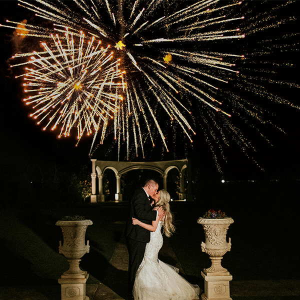 wedding-fireworks-classic Bride and groom share a romantic kiss beneath vibrant fireworks in front of a picturesque garden archway at night. | Sky Rye Design Bride and groom share a romantic kiss beneath vibrant fireworks in front of a picturesque garden archway at night.