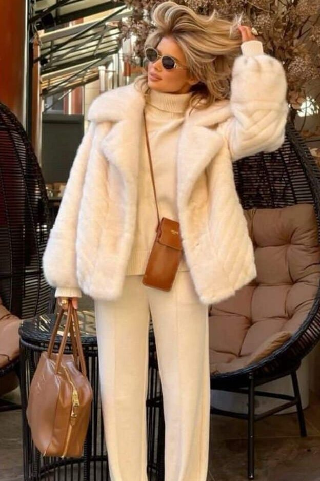 Fashionable woman in a cozy white outfit, sunglasses, and a fur coat, holding a brown bag, exuding chic winter style.