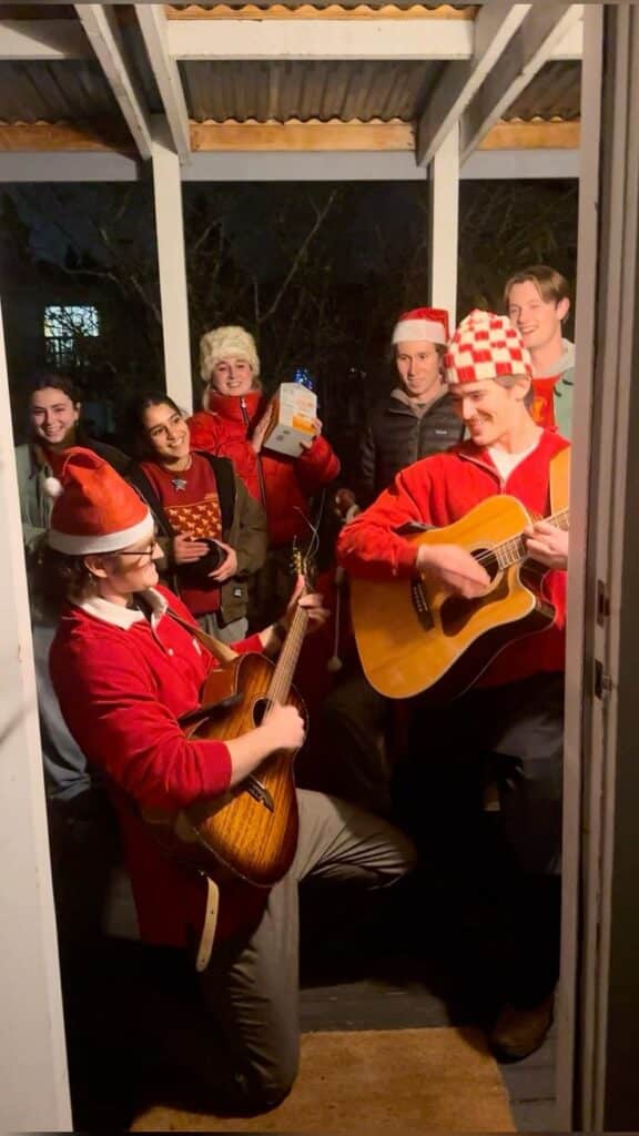 Group of people in festive hats caroling on a porch, two playing guitars, spreading holiday cheer at night.