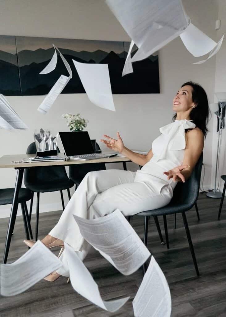 Professional woman joyfully throws papers in office, celebrating success with laptop and awards on desk.