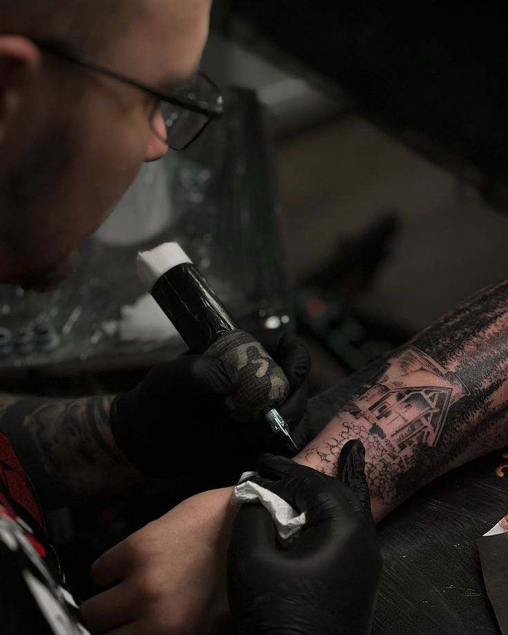 Процессы в студии Tattoo artist in black gloves works on an intricate arm design featuring a house and trees, using a tattoo machine. | Sky Rye Design Tattoo artist in black gloves works on an intricate arm design featuring a house and trees, using a tattoo machine.