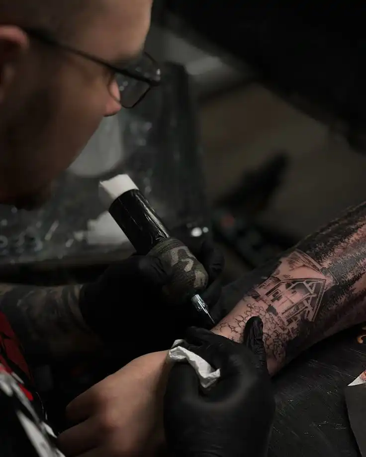 Tattoo artist in black gloves works on an intricate arm design featuring a house and trees, using a tattoo machine.