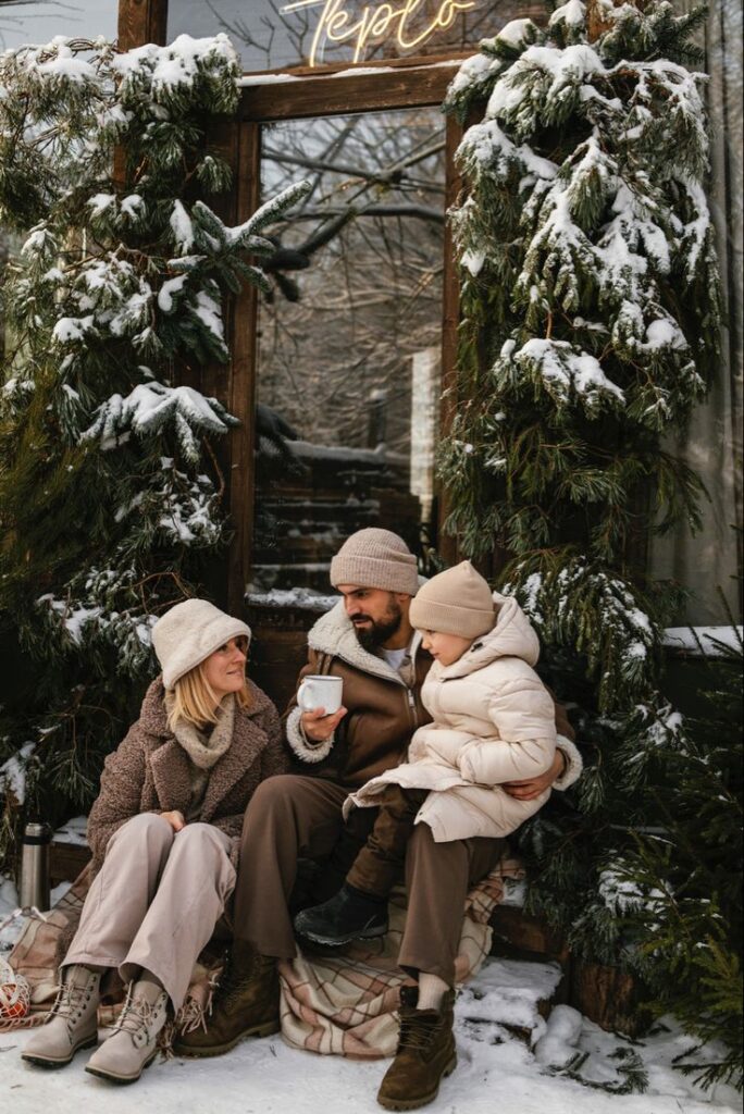Семейная фотосессия Family in winter clothing enjoying hot drinks outside a snowy cabin decorated with fir branches, creating a cozy winter scene. | Sky Rye Design Family in winter clothing enjoying hot drinks outside a snowy cabin decorated with fir branches, creating a cozy winter scene.