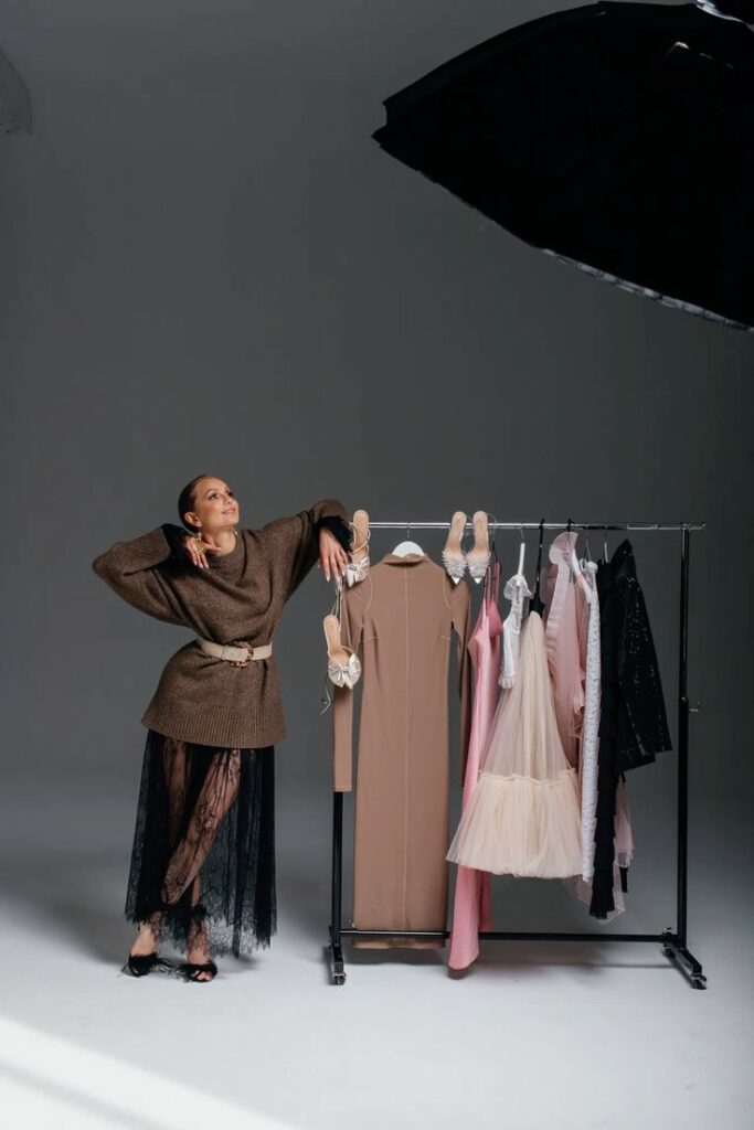 Fashionable woman in a brown sweater poses beside a clothing rack with elegant dresses in a photo studio setting.