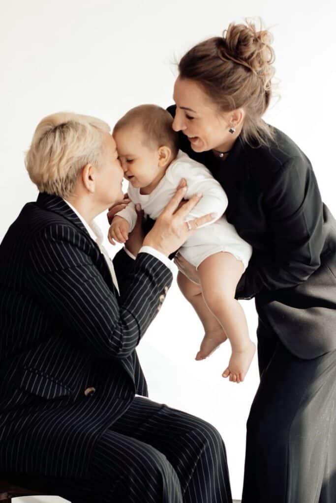 Три поколения фотосесси в стиле минимализм Three people sharing a joyful moment: an adult in a pinstripe suit kisses a smiling baby held by another adult in a black outfit. | Sky Rye Design Three people sharing a joyful moment: an adult in a pinstripe suit kisses a smiling baby held by another adult in a black outfit.