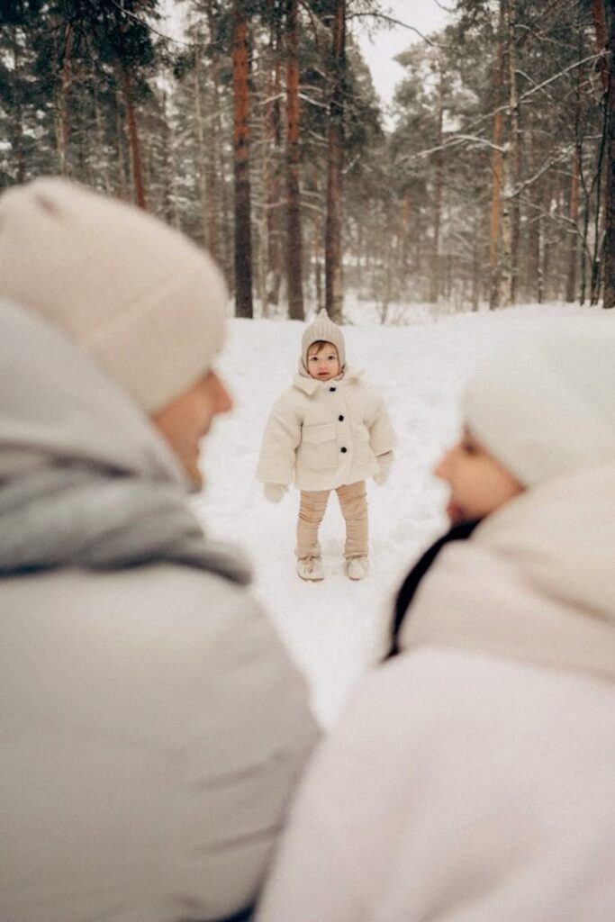 Фотограф Мария Струтинская_ strutinskayamariya Child in a white winter coat stands in snowy forest, facing parents in the foreground. Trees in the background covered with snow. | Sky Rye Design Child in a white winter coat stands in snowy forest, facing parents in the foreground. Trees in the background covered with snow.