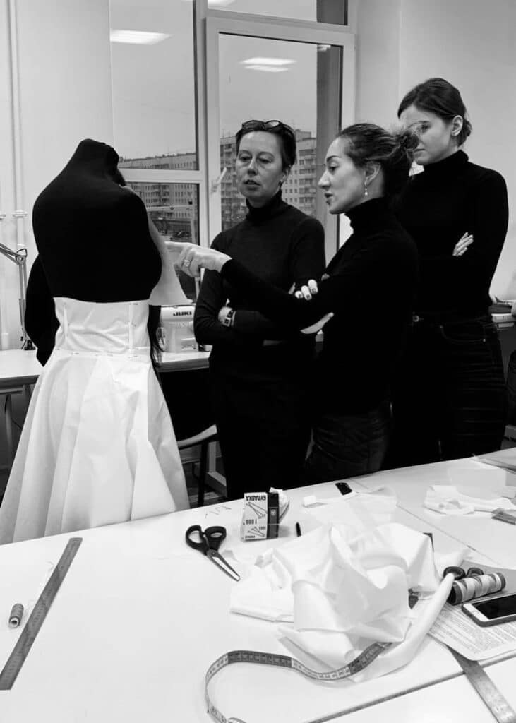 Fashion designers collaborate on a white dress in a studio, surrounded by sewing tools.