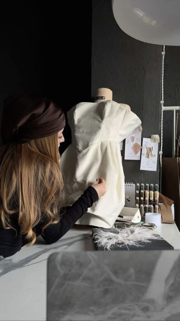 Screenshot Fashion designer sewing a white dress on a mannequin in a stylish studio, surrounded by sewing materials and design sketches. | Sky Rye Design Fashion designer sewing a white dress on a mannequin in a stylish studio, surrounded by sewing materials and design sketches.