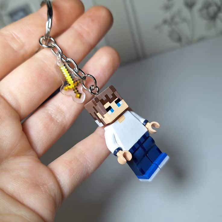 брелок лего майнкрафт Close-up of a hand holding a Minecraft-inspired character keychain with a small yellow sword charm attached. | Sky Rye Design Close-up of a hand holding a Minecraft-inspired character keychain with a small yellow sword charm attached.