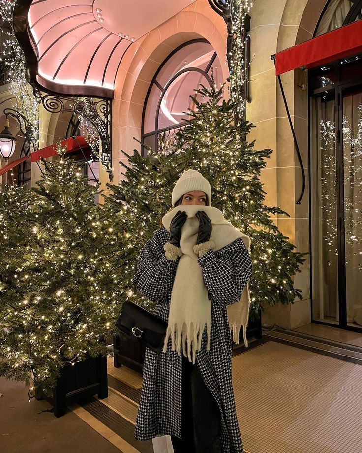⚡️🤍 Person bundled up in winter attire stands in front of festive, decorated Christmas trees outside elegant building entrance. | Sky Rye Design Person bundled up in winter attire stands in front of festive, decorated Christmas trees outside elegant building entrance.