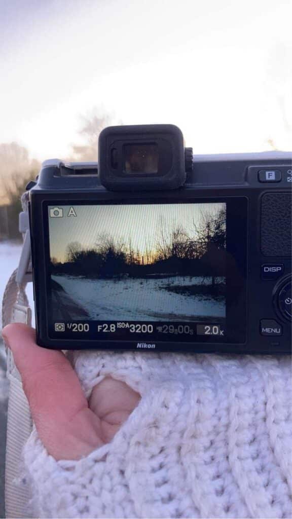 ❄️Hiie❄️ Camera displaying a snowy landscape with trees at sunset, held by a hand in a cozy sweater. | Sky Rye Design Camera displaying a snowy landscape with trees at sunset, held by a hand in a cozy sweater.