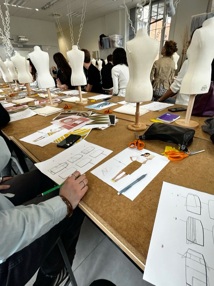 🧵🪡 Fashion design students in class, sketching and working at a table with mannequins and design drawings, scissors, and tools visible. | Sky Rye Design Fashion design students in class, sketching and working at a table with mannequins and design drawings, scissors, and tools visible.