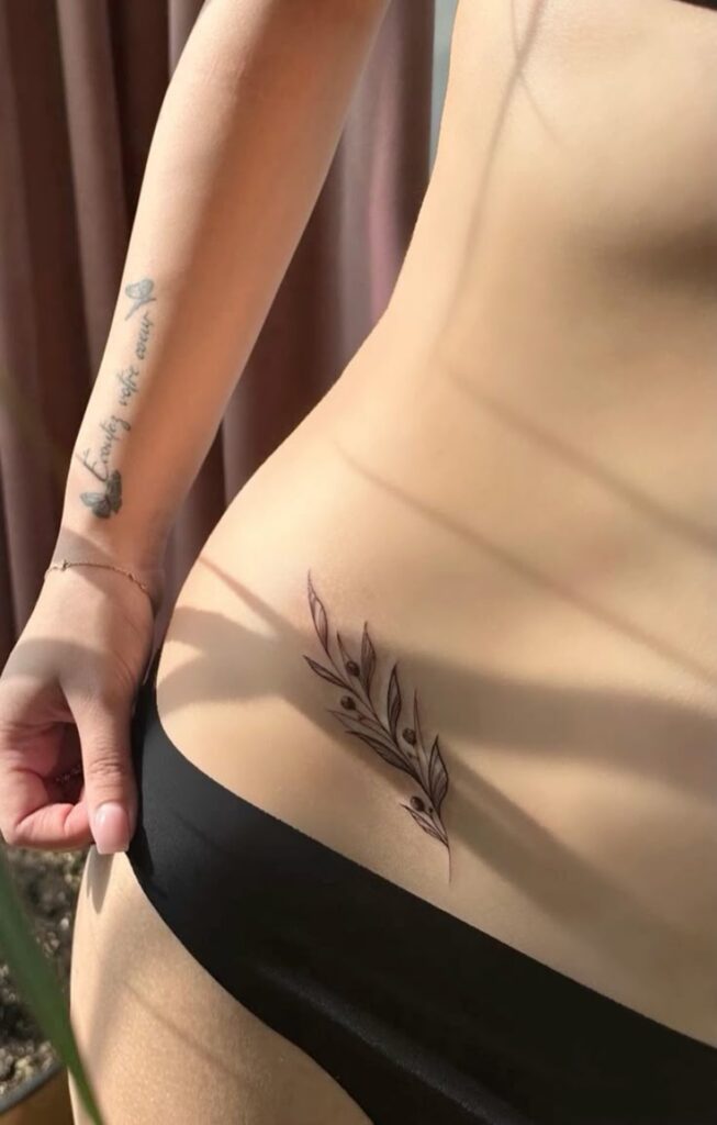 Татуировка на бедре Minimalistic leaf tattoo on lower hip, complemented by a quote tattoo on arm, in natural light. | Sky Rye Design Minimalistic leaf tattoo on lower hip, complemented by a quote tattoo on arm, in natural light.