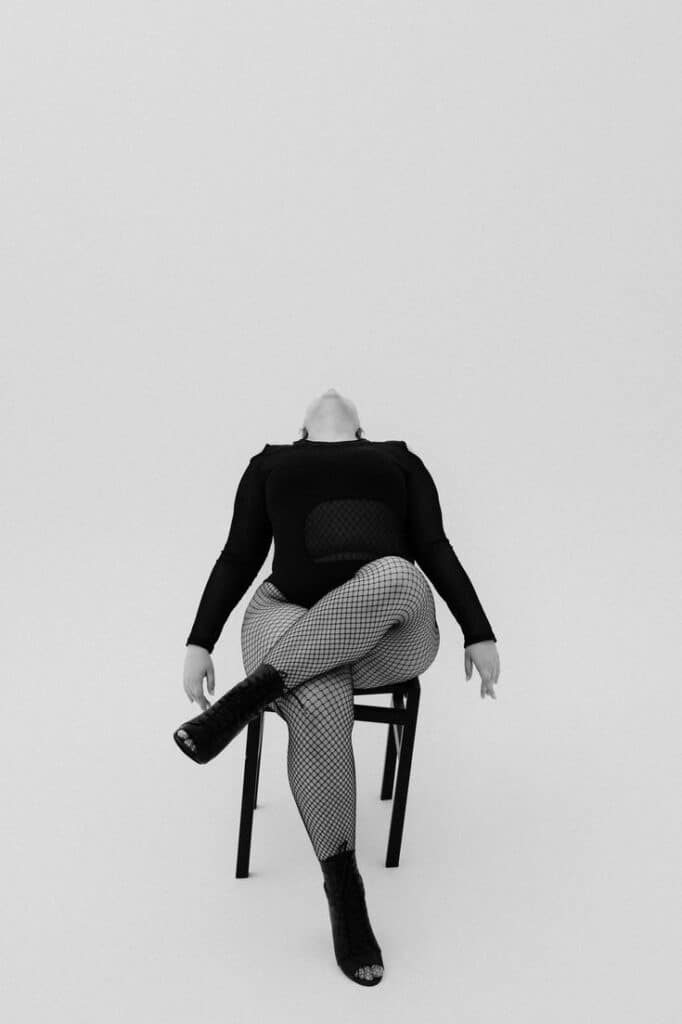 зйомка plussize curves style Person in stylish black outfit with fishnet tights and boots, posed on a chair against a minimalist background. | Sky Rye Design Person in stylish black outfit with fishnet tights and boots, posed on a chair against a minimalist background.