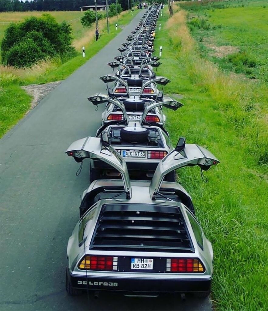 A line of DeLorean cars with gullwing doors open on a country road, showcasing iconic automotive design.