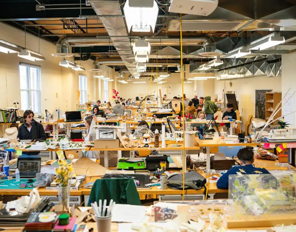 Busy design studio with people working on architectural projects, surrounded by desks, computers, and creative materials.