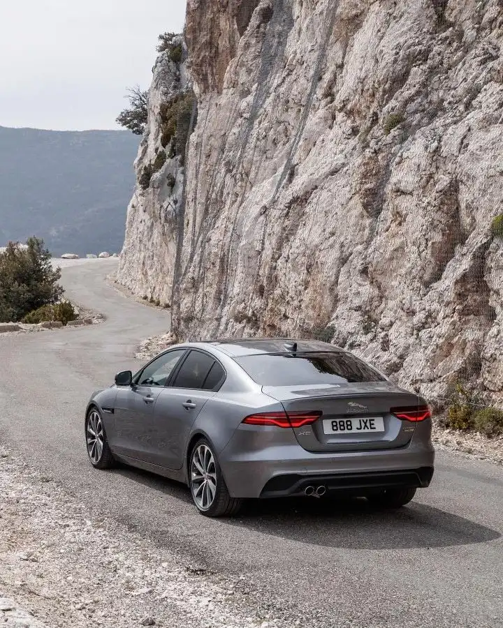 Luxury silver sedan driving on winding mountain road with rocky cliffs, showcasing sleek design and performance.