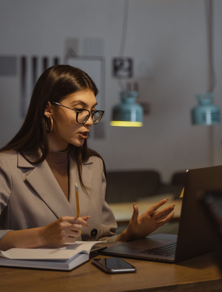 _ 1 Woman in glasses working on a laptop, holds a pencil, and writes in a notebook at a cozy workspace with pendant lighting. | Sky Rye Design Woman in glasses working on a laptop, holds a pencil, and writes in a notebook at a cozy workspace with pendant lighting.